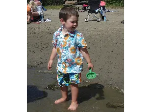 Toddler in a flowered shirt playing on a lakeshore with a sand shovel